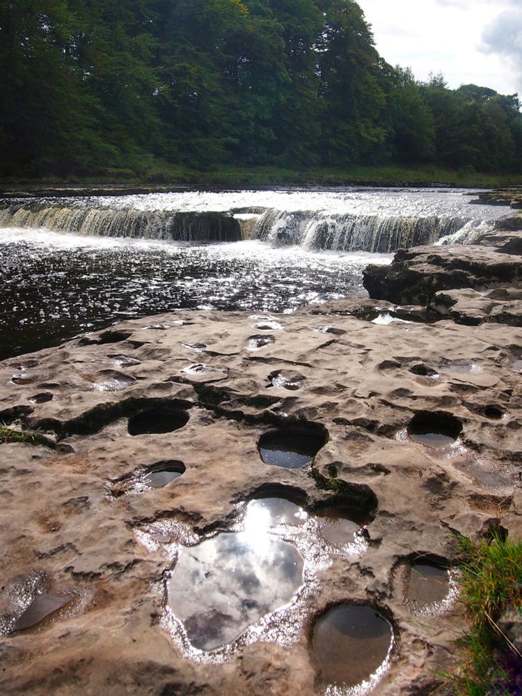 Mit vielen, teils wassergefüllten Löchern übersätes steinernes Ufer eines Flusses. In der Mitte eine kleine Stufe, über die das Wasser fällt, im Hintergrund Wald.