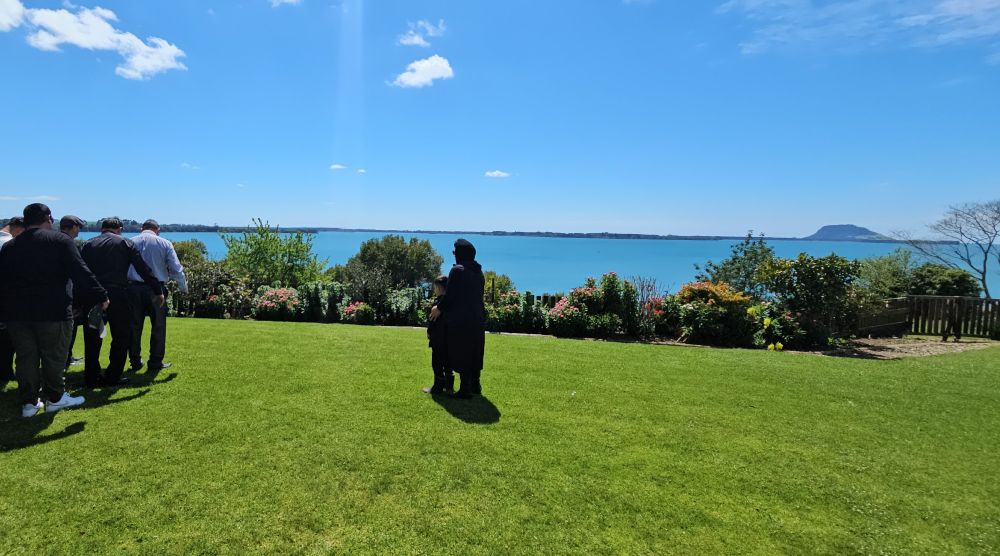 Women (centre) in black standing in a yard with a child in front of her watching as men are standing looking solemn in a group (left of image) Backdrop is Tauranga harbour looking out to Mauao, Matakana on a sunny day