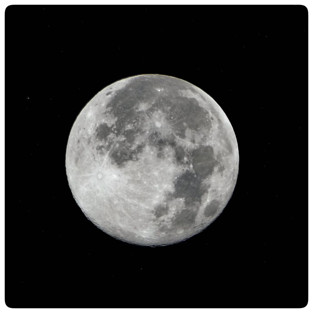 The image depicts a detailed view of the November 5, 2024 supermoon (also known as Beaver Moon) against a black background. The supermoon is the primary subject, dominating the center of the frame. Its surface is textured with numerous craters, ridges, and dark patches, giving it a rough, three-dimensional appearance. The colors are primarily shades of gray and white, with the darker areas representing the lunar "seas" or lowlands and the lighter areas highlighting the highlands and impact craters.

The background is a deep black, punctuated by small, bright stars. These stars are scattered randomly, adding depth and a sense of vastness to the scene. The image is framed within a white border, which creates a sense of separation and focus. The overall composition is symmetrical, with the full moon centered, drawing the viewer's eye directly to its detailed surface. The image evokes a sense of wonder and the mystery of space.