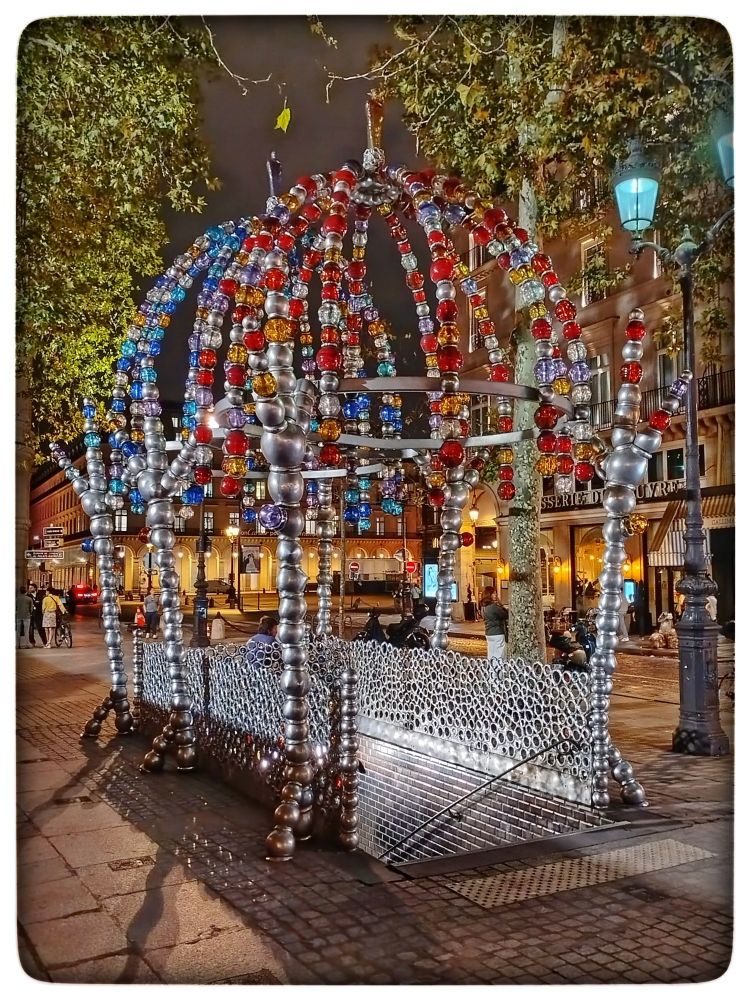 Palais Royal - Musée du Louvre metro exit known as the "Kiosque des Noctambules" on Place Colette in 75001 Paris, France.
Inspired by Murano glass and Venetian master glassmakers, artist Jean-Michel Othoniel designed an enchanting kiosk that contrasts with the classical architecture of the Parisian square.
The "Kiosque des Noctambules" consists of two gloriette domes, a veritable explosion of color:
The first dome, adorned with warm-toned pearls, evokes daylight.
The second dome, adorned with cool-toned pearls, represents night.
Topped by glass statuettes made by the famous Salviati glassworks, these two domes offer a striking visual spectacle.
The kiosk sparkles with a thousand lights when illuminated by the sun or public lighting. The experience continues inside the metro station, where gilded tiles, glass beads and a public bench add to the enchanting atmosphere.
The Palais Royal - Musée du Louvre station thus becomes more than just a place of transit. It's an invitation to look at the world through the eyes of a child, a celebration of the joy that colors our daily lives.