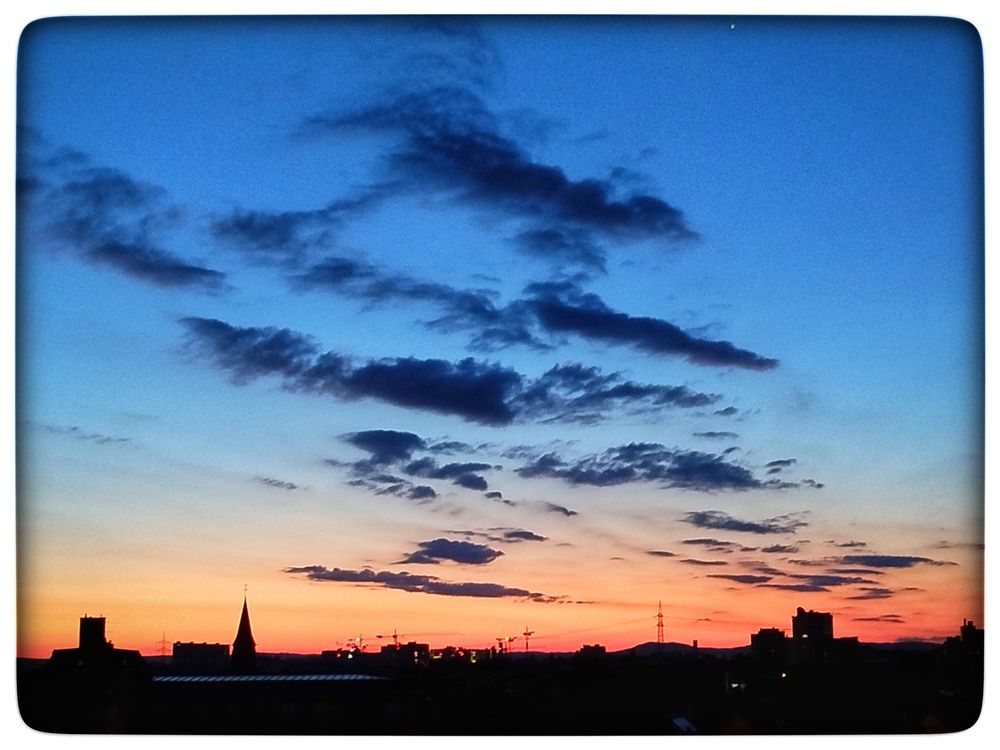 The image captures a vibrant sunrise over a city skyline. The sky is the dominant element, showcasing a gradient of colors. The upper portion of the sky transitions from a deep, rich blue to a lighter, almost pastel blue. Scattered across this blue canvas are dark, textured clouds, adding depth and visual interest.

Below the blue, the sky transforms into a warm palette of orange and red, indicating the sun's position below the horizon. The hues blend smoothly, creating a soft and inviting glow.

The cityscape is silhouetted against this colorful backdrop. Buildings of varying heights and shapes are visible, including what appears to be a church steeple, which adds a distinct architectural element to the scene. Construction cranes are also visible, hinting at ongoing development within the city. The buildings and structures are rendered in dark tones, emphasizing their forms against the bright sky.

The composition is framed with a rounded border, which gives the image a slightly softened and nostalgic feel. The overall impression is one of tranquility and beauty, capturing a fleeting moment of natural splendor over an urban landscape.