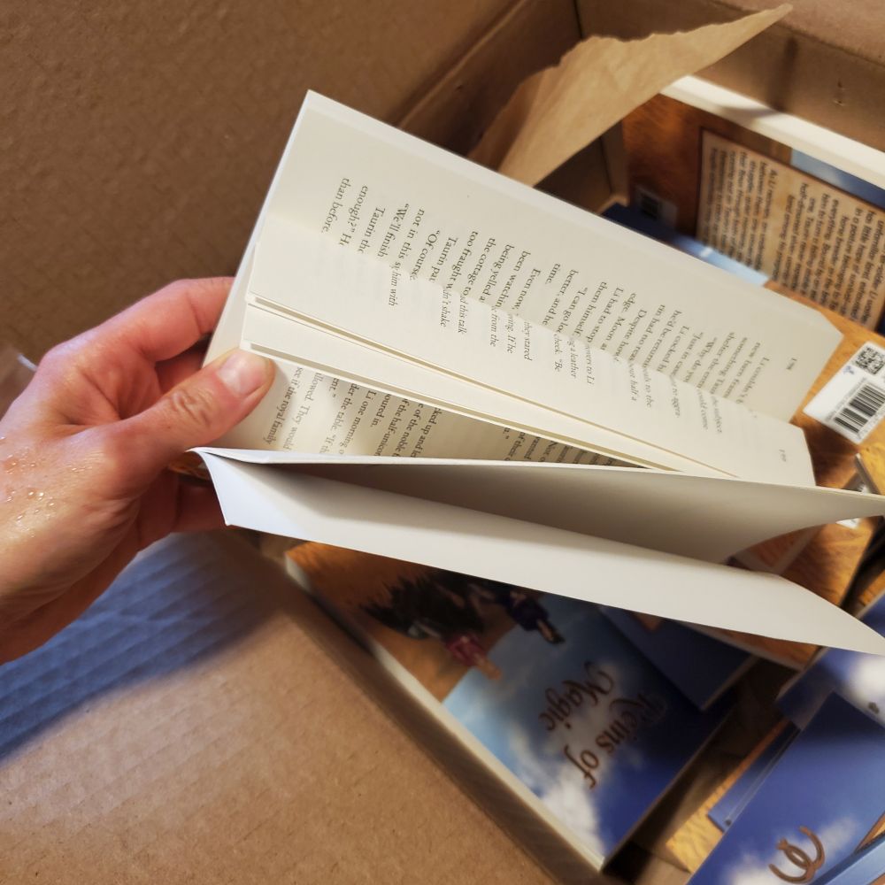 Photo of a hand holding a paperback copy of Reins of Magic with some of the pages bent in half due to improper packaging.