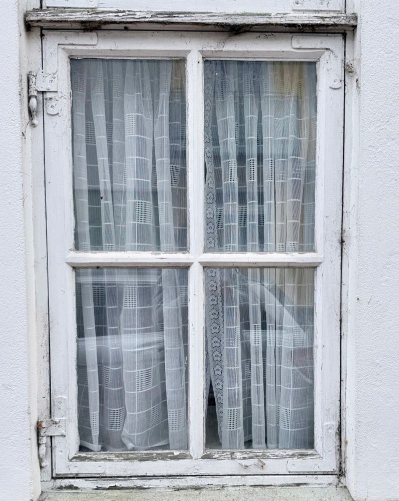 Weißer Fensterrahmen, die Farbe teilweise abgeblättert. Im Fenster eine Gardine und in den Scheiben spiegelt sich ein parkendes Auto. 