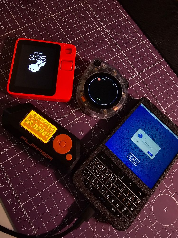 A photo of various electronic devices. The setup includes a red handheld device with a bunny icon on the screen, Rabbit R1, a circular transparent gadget with visible internal components, my OrionKit, a black and orange Flipper Zero displaying "Mr. Robot" on its screen, and a custom handheld device running Kali Linux with a keyboard and touchpad, my HackberryPi