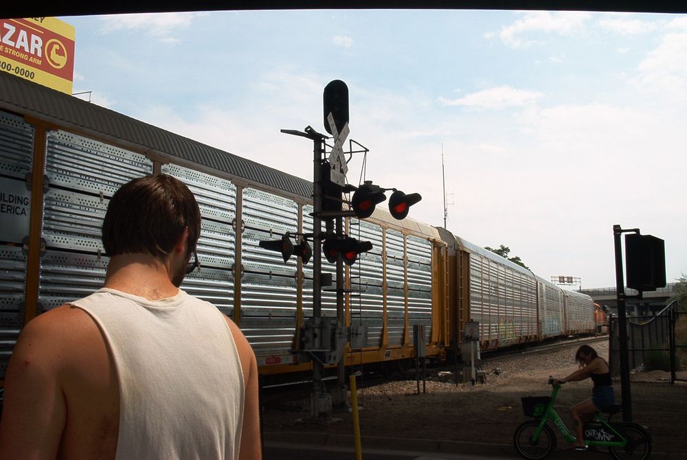 picture of a person in a white tank top standing in front of a railroad crossing, with a train of yellow and silver car carriers on the tracks 