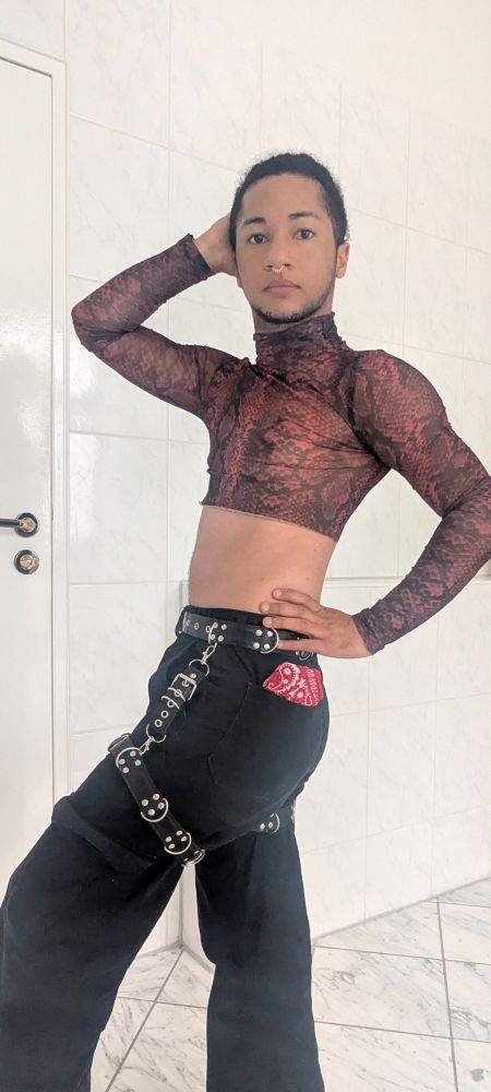 Glitx, a mix transmasc person, is posing for the camera. He's wearing a mesh crop top with turtle neck, black pants with a latex harness on his thighs and waist, and there's a red handkerchief in his left pocket. 