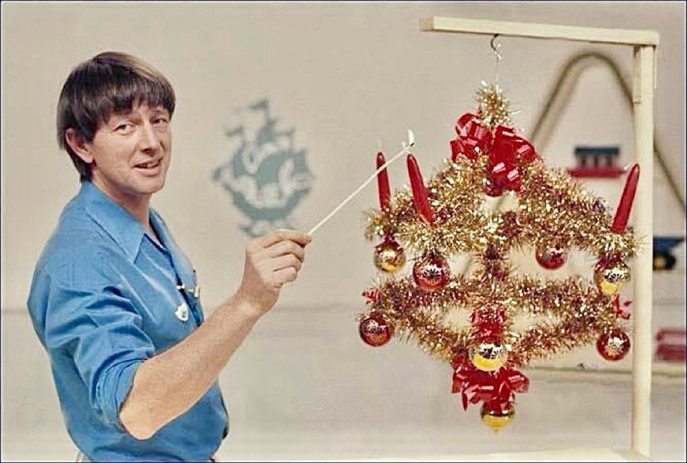 BBC Archive photo - Blue Peter studio - John Noakes holding a lit taper as he lights the first red candle on an Advent crown made from coat hangers covered in gold tinsel. The coat hangers are suspended from a spindly wooden gallows frame.
