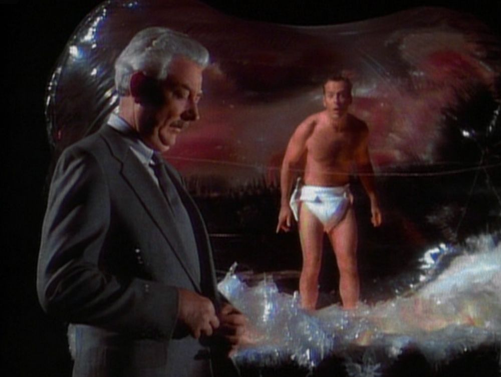 Actor Bruce Willis wears a diaper and stands inside a clear inflated structure representing a uterus. A dapper man in a grey suit stands outside the uterus.