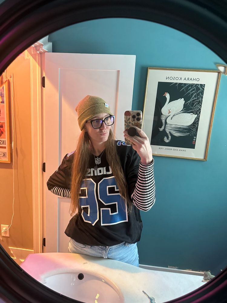 Image is a woman with long hair taking a selfie in a mirror while wearing a Detroit Lions jersey. 