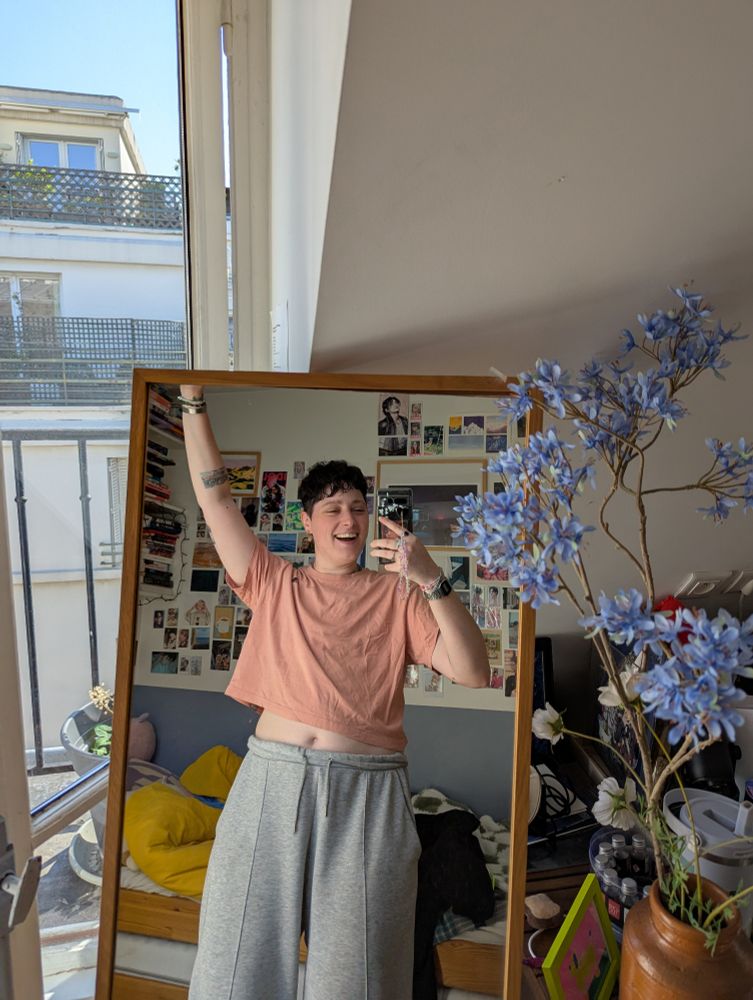 photo de moi dans un miroir, je porte un legging gris et un t-shirt un peu cropped rose saumon et je souris la bouche ouverte avec un bras en l'air pour signifier ma joie 