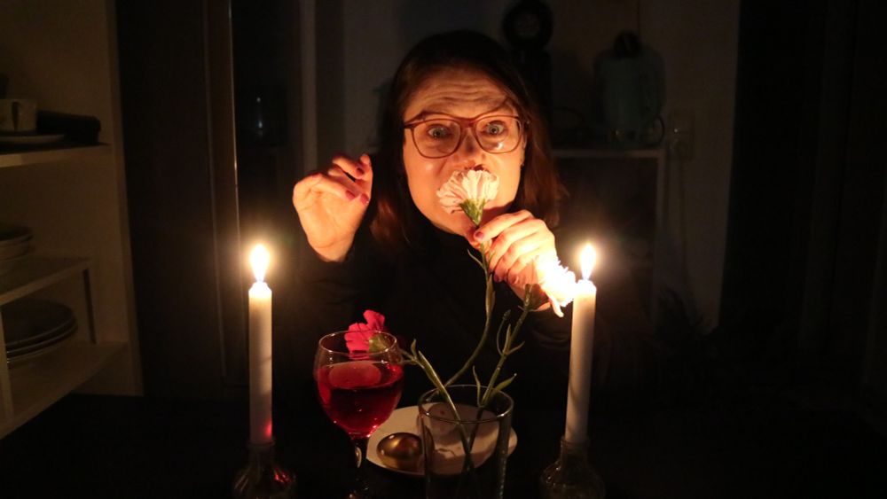 Lara (Frau) sitzt an einem gedeckten Tisch im Dunklem beleuchtet durch nur zwei Kerzen und beißt in die Plastikblume während sie in die Kamera schaut 