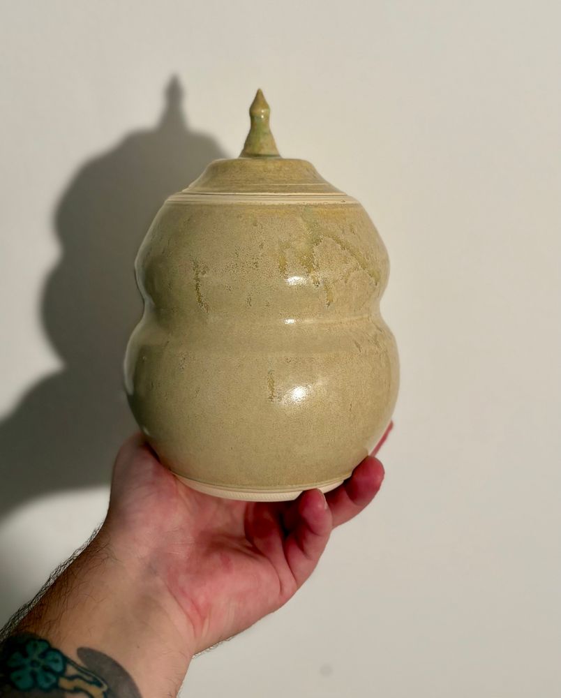 A hand holds up a tan ceramic jar against a white background. The stout jar has a slight waist, and the knob of the lid is tall and pointed. It features a lightly variegated tan glaze splattered with nearly translucent green spots.  