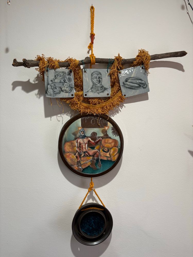 3 hand drawn, blue porcelain tiles hang from a poplar branch. The leftmost features a room with two chairs, the middle a portrait of a man wearing a striped balaclava with a heart shaped face opening, the third a hotdog with a price Albert piercing. Below is a painted disk featuring an oil painted tableau  featuring the 3 elements of the tiles combined: a man wearing a striped balaclava sitting on the chairs in the room, with the pierced hotdog  on the chairs next to him. Below that is a disk with an inset of carpet that matches the painted scene. All these elements are hanging on a yellow rope. 