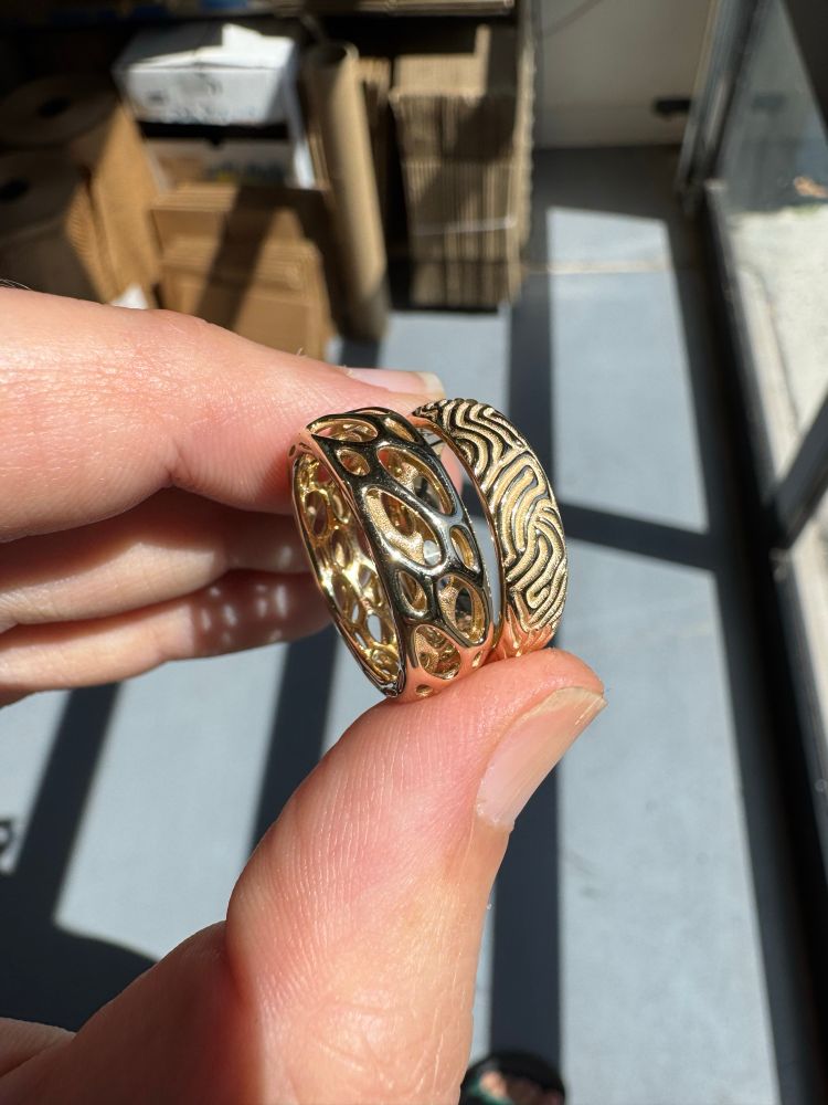 Two 14k yellow gold rings in my fingers. The left one is a cellular skeleton with two layers of cells twisting around the ring. On the right a reaction diffusion ring with sand dune like ridges and valleys