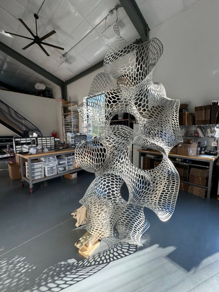 Photo of the inside of our studio it’s a two-story high room with white walls and a white ceiling and a giant aluminum biomorphic undulating cellularly perforated sculpture standing in the middle
