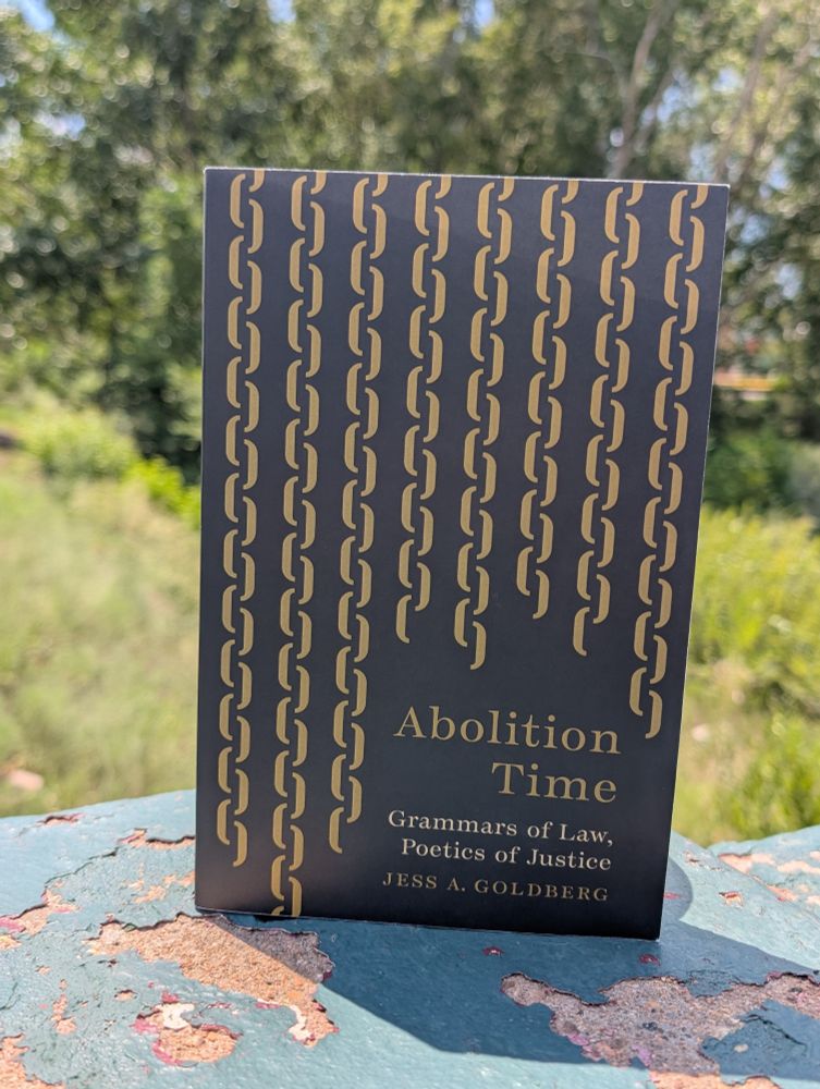 A copy of the book "Abolition Time: Grammars of Law, Poetics of Justice" is shown propped up on a ledge against a background of trees and greenery.