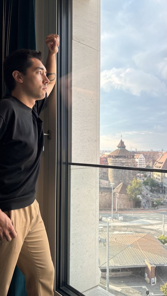 A person stands by a large window, looking out at a cityscape featuring historical buildings and a cloudy sky.