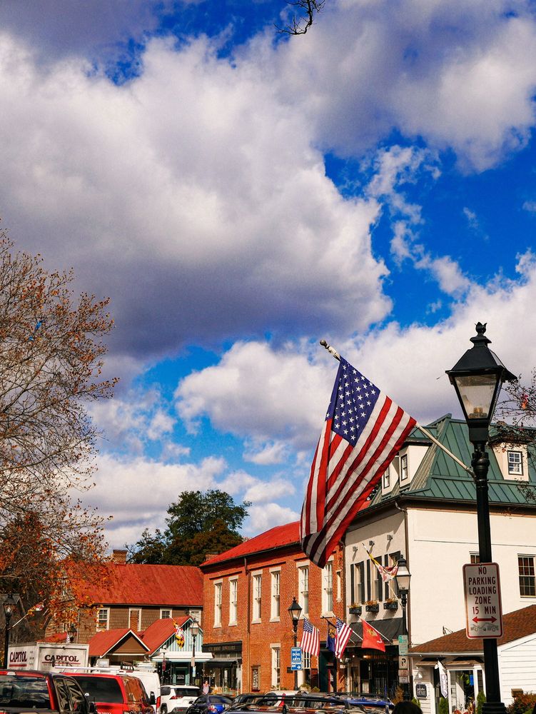 A busy small-town street features American flags, historic-style buildings, and a bright, partly cloudy sky.