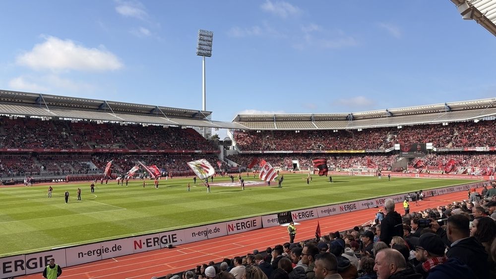 A large crowd at a bustling soccer stadium watches as players and staff display flags on the field with vibrant banners in the stands.