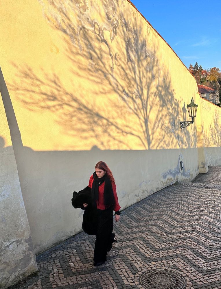 Redhead girl wearing red sweater walks uphill towards Prague castle.  The late afternoon sun casts shadows of trees against the sun on wall. The yellow stands out against blue of the sky.