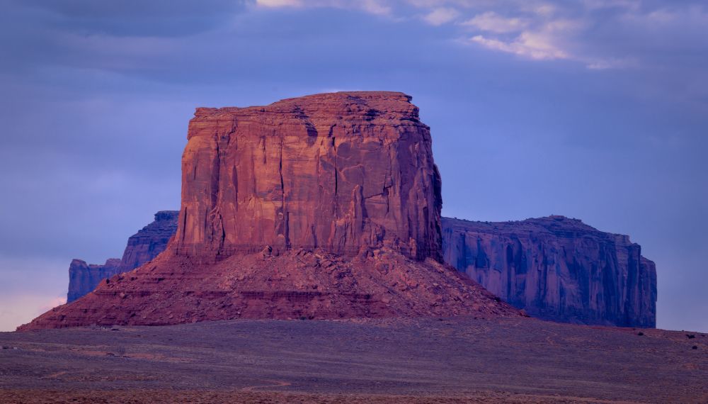 A striking landscape featuring a tall, rugged red rock formation under a twilight sky with hues of purple and blue. The formation is surrounded by a flat, barren terrain with additional rock structures in the background, captured during a serene evening.

Photography by: David Lennard ©2025