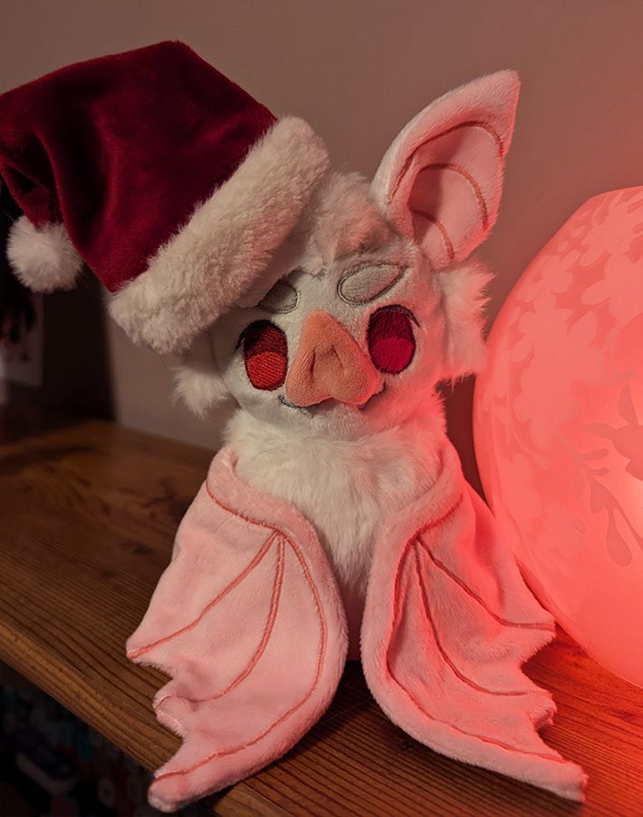 A plush of batstarion wearing a small santa hat, illuminated by a pink lamp.