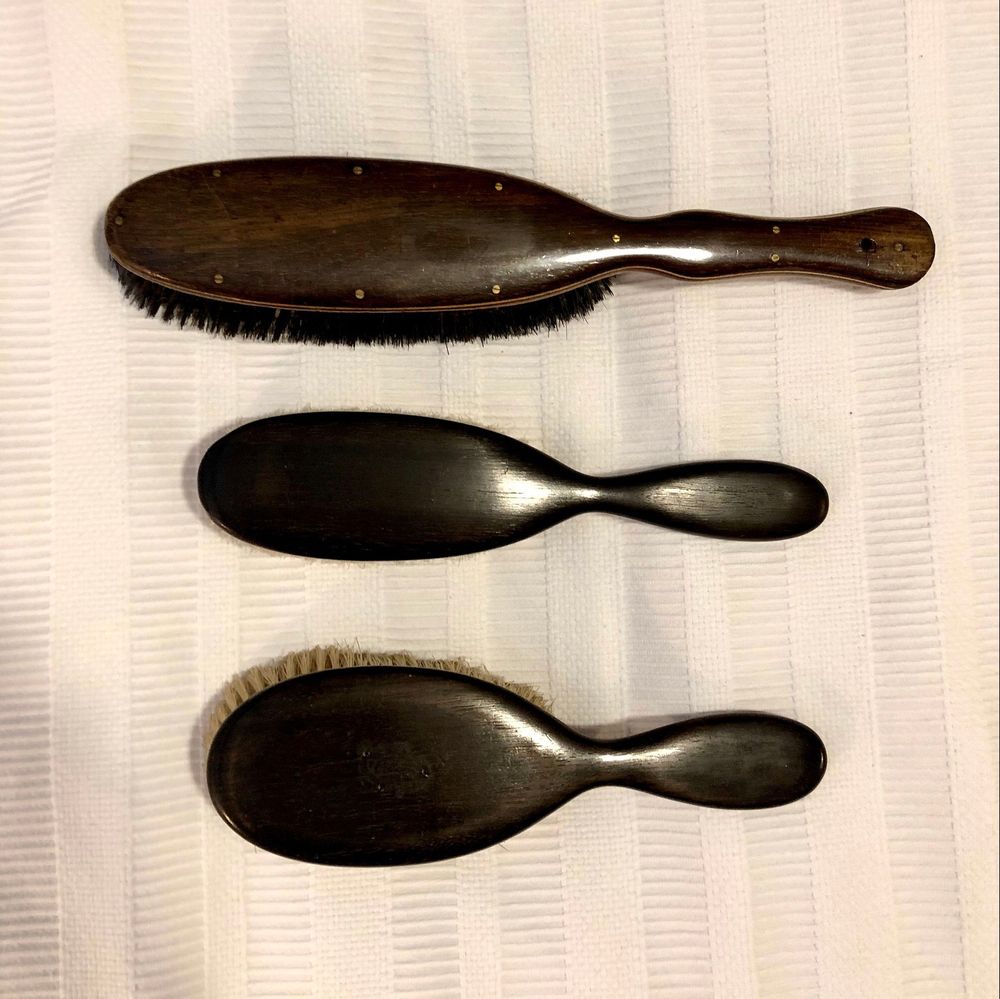 A photo of three brushes from my spanking implement collection. Top to bottom: a long brown wooden (unknown type) clothes brush with nine small flush brass screws around the edges and along the handle; a long, narrow ebony hairbrush; and a broader/rounder more traditionally-shaped ebony hairbrush,