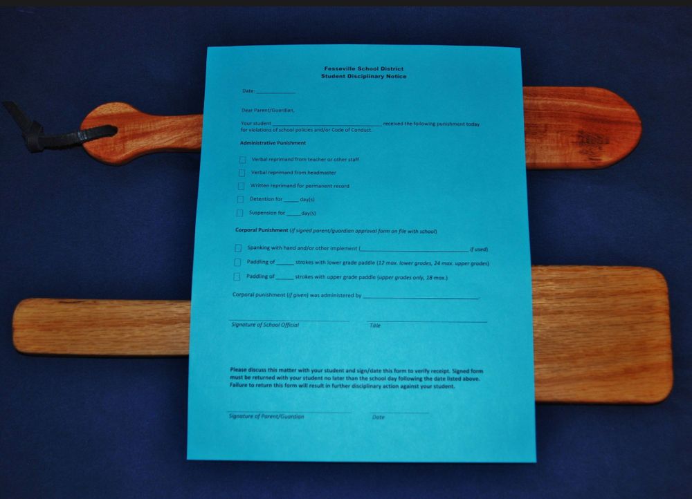 A light blue sheet of paper with a fictional form entitled “Fesseville School District Student Disciplinary Notice” sitting on top of two wooden paddles