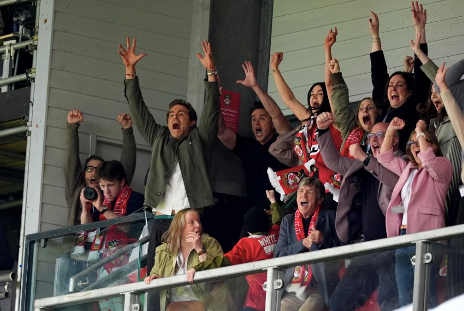 A packed director's box at Wrexham football ground. The majority of the people, including Ryan Reynolds and Rob McElhenney have their hands high in the air in celebration. At the front of the box is Archie White who has turned towards Ryan and Rob. He is wearing a black beanie hat and a red Wrexham football shirt with White written on the back. His mother is seated beside him and looks on in shock and wonder.