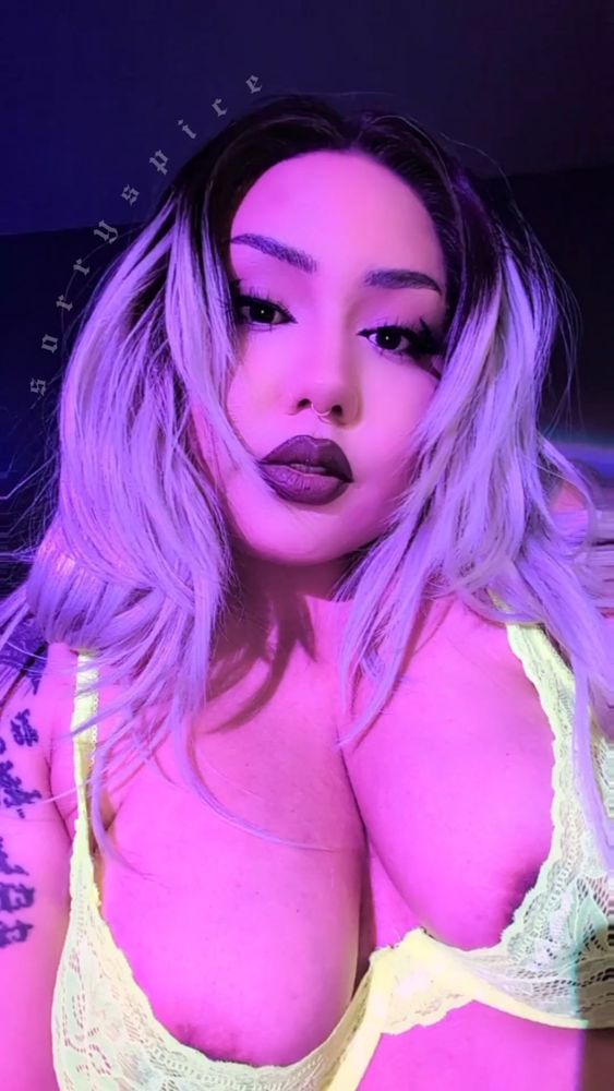 Spice is wearing a neon green bralette that glows yellow in their pink/purple sunset lamp. Camera angle is low so their head is slightly tilted towards it, staring you down. They have long blonde hair that looks pink/purple in the lighting. Sharp eyeliner, dark brown lipstick, and facial piercings. 