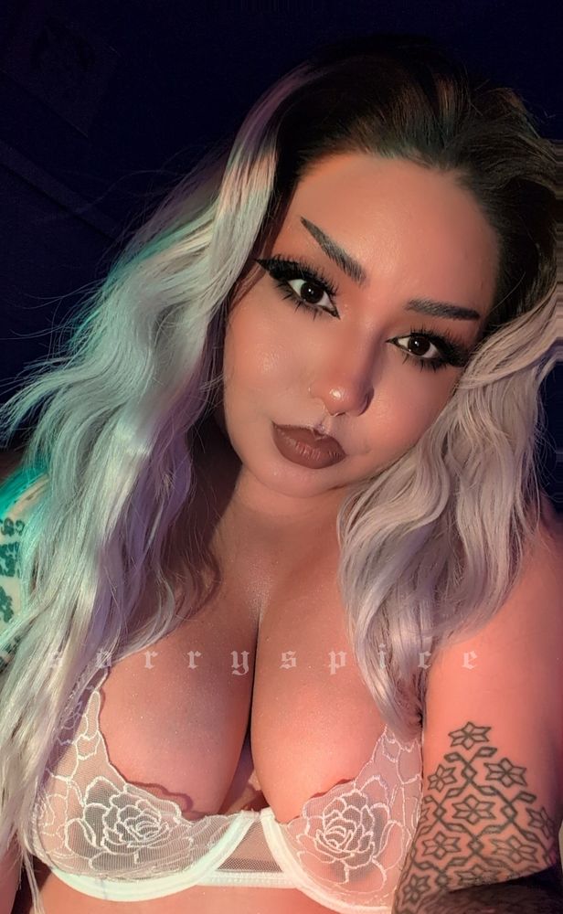 Spice with long blonde hair in a lacey white bralette staring lovingly into the camera. They have tattoos on both arms, sharp eyeliner, and kissable soft brown lips 