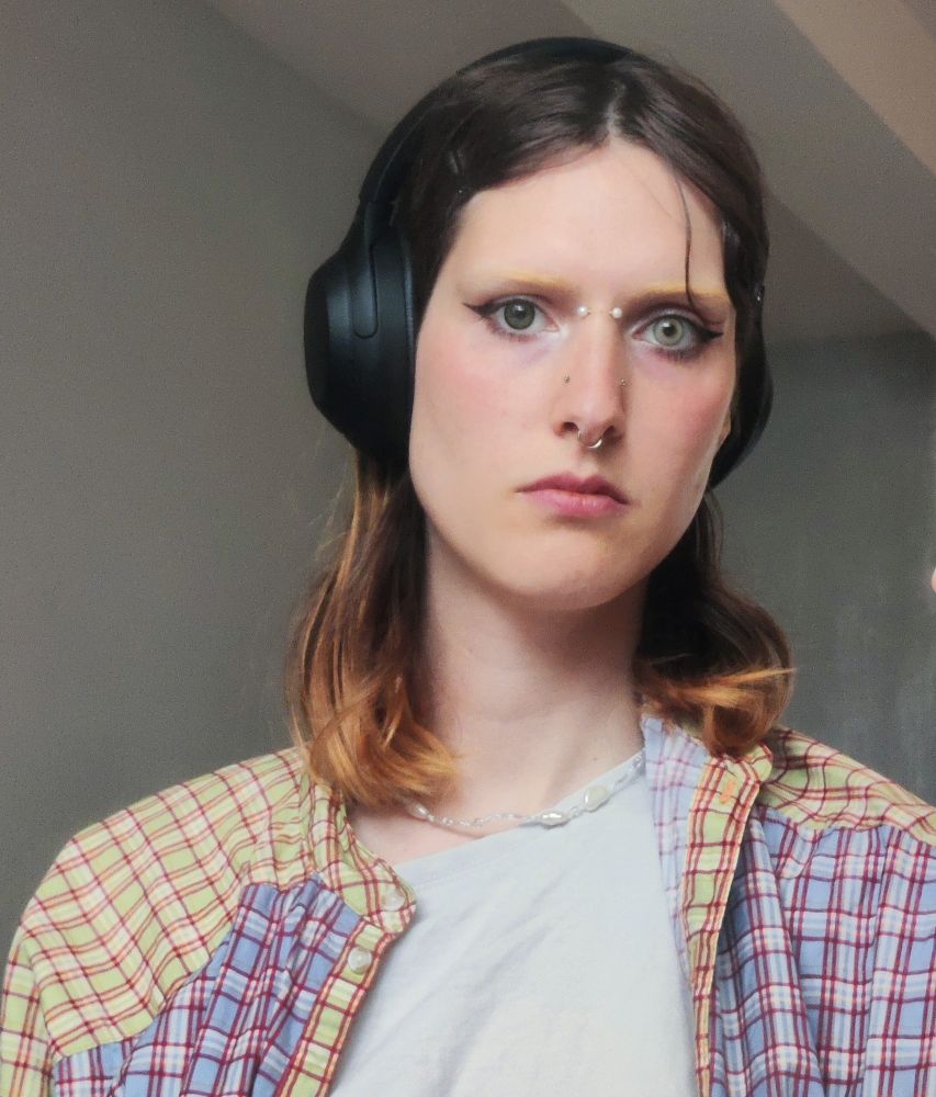 selfie of a woman with light blush and eyeliner. she is wearing colored contacts, with the right eye being a light gray and the right one a darker emerald green. she has bleached eyebrows and shoulder length brown hair. she is wearing headphones, covering one ear only. she is wearing an open checkered shirt over a white t-shirt, and a pearl necklace. she has a bridge piercing with pearl barbell balls, small golden high nostril piercings, and a gold ring septum. 