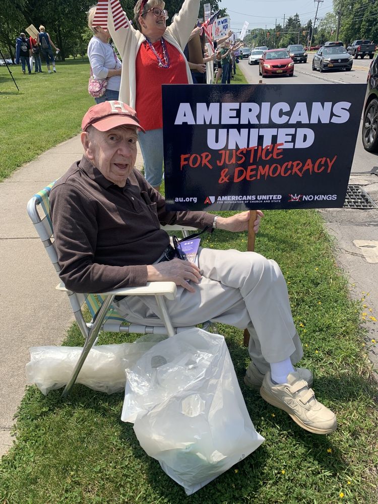 image of a 92 year old man sitting in a lawn chair holding up a sign that says Americans United for Justice and Democracy