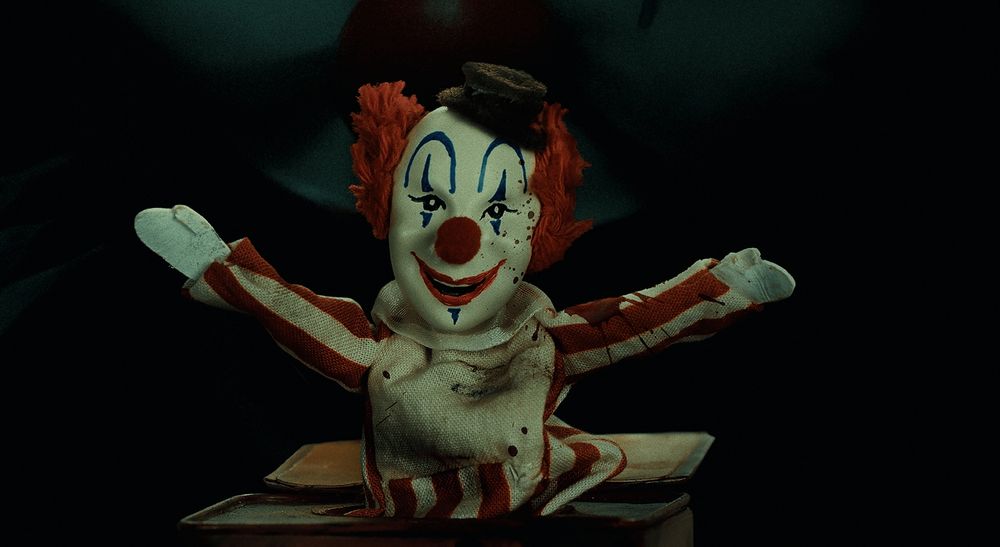 A creepy toy of Frendo the clown popping out of a music box with his arms spread wide. Oh also there's a much more real clown face lurking in the darkness behind it