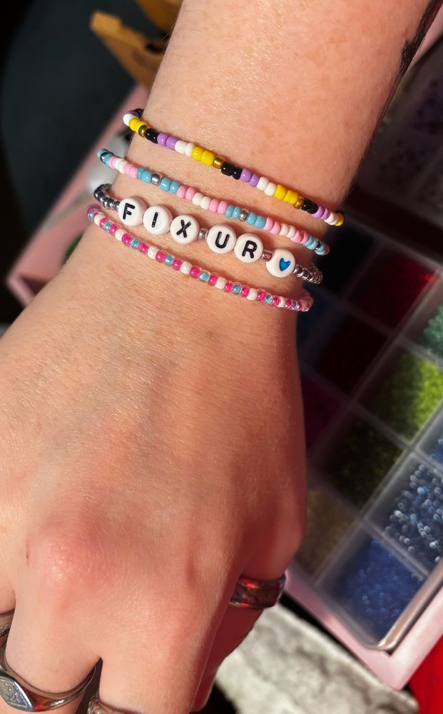Georgie’s hand with bracelets she has made in the colours of the trans pride flag, the nonbinary flag, and one that reads “fix ur heart”