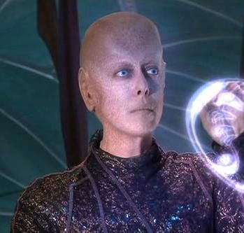 Da'an from Earth: Final Conflict. A being deliberately portrayed as genderless, and canonically semi energy based rather than matter based like humans. They do however have a more solid form as seen in this image pale skin, slightly molted in colour. No hair. Large flat ears pressed against their side. Pale blue eyes and a large forehead in a way that looks graceful. Wearing a slightly shiny purple body suit. 