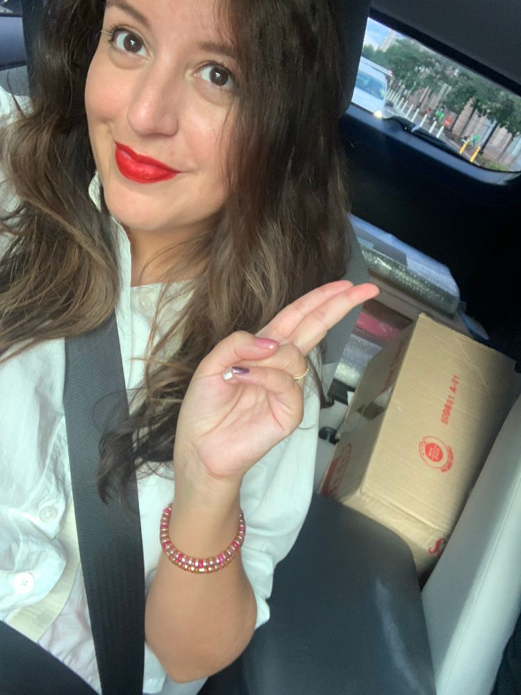 Young woman with long brown hair sits on car’s passenger seat, seatbelt on, holding two fingers up to make an ancient peace sign. The back of the car holds different packaged artworks. 