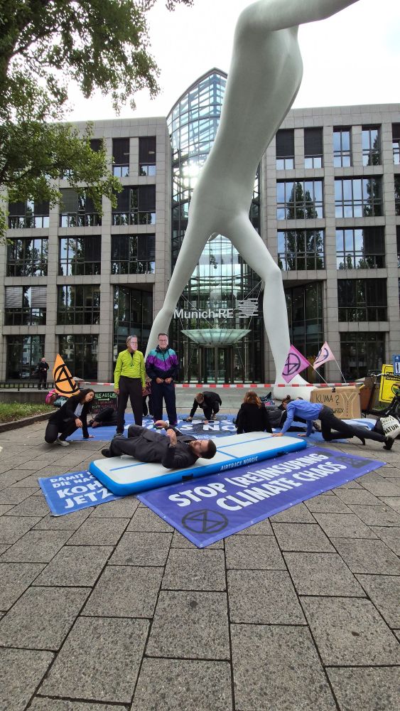 Aktivisti vor der Zentrale der Munich RE in München. Verkleidet als CEO Wenning u. Jurecka in Trainingsanzügen. Ein als Mitarbeiter verkleideter Aktivist übt auf einer Turnmatte die Rolle Rückwärts