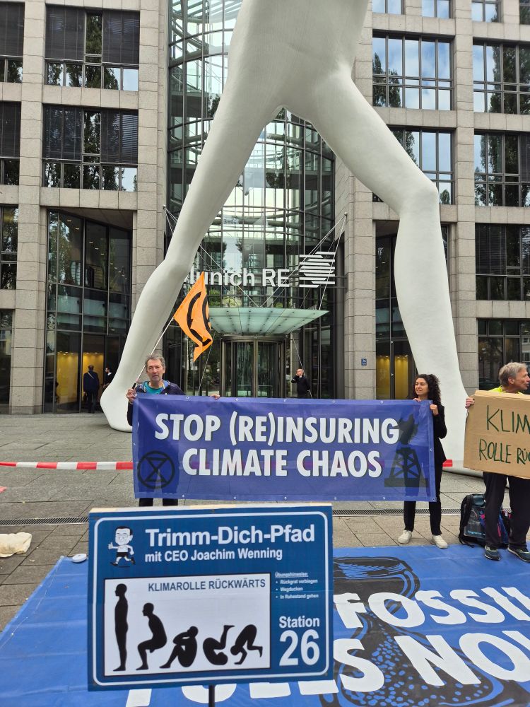 Aktivisti vor der Zentrale der Munich RE in München. Ein Schild macht auf den aufgebauten Trimm-Dich-Pfad aufmerksam mit Anleitung für die Rolle Rückwärts.