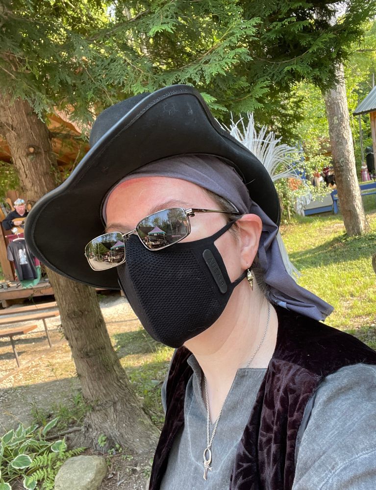 A pale-skinned human wearing sunglasses, a black face mask, a gray bandanna, and a pirate hat takes a selfie in front of some trees at a Renaissance Festival