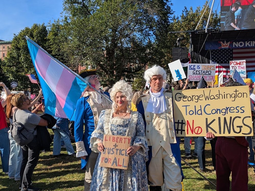 A trio of people dressed in historic clothing. There are two men in the back dressed as George Washington and the woman in front is dressed as Martha Washington. She holds a sign that says Liberty and justice for all. The George on the left holds a Trans pride flag which is blue, pink and white striped. The George on the right holds a sign that says George Washington Told Congress #NoKings