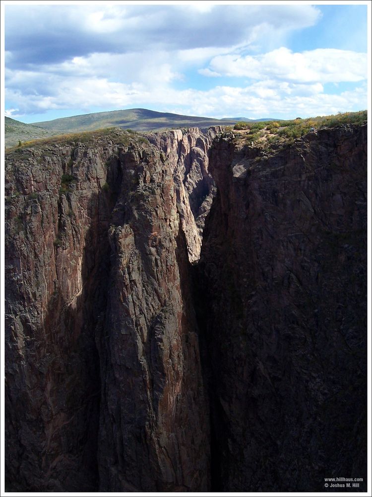 A landscape photo of the Black Canyon of the Gunnison. 2/3 of the photo is below land, showing a steep decline into an abyss below rolling hills and slightly cloudy blue sky.