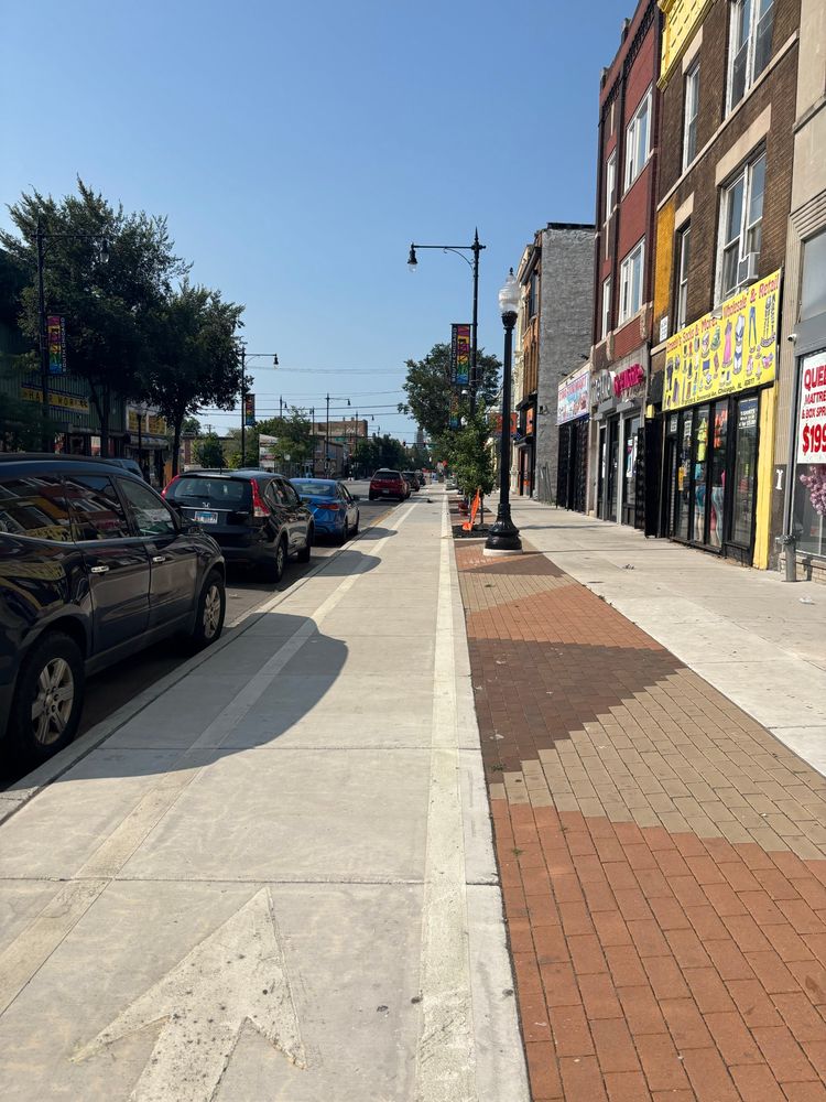 Sidewalk height bike lane on S Commercial Ave in South Chicago. Parked cars to the left and sidewalk+storefronts on the right. Patterned brown bricks delineate sidewalk space from bike lane