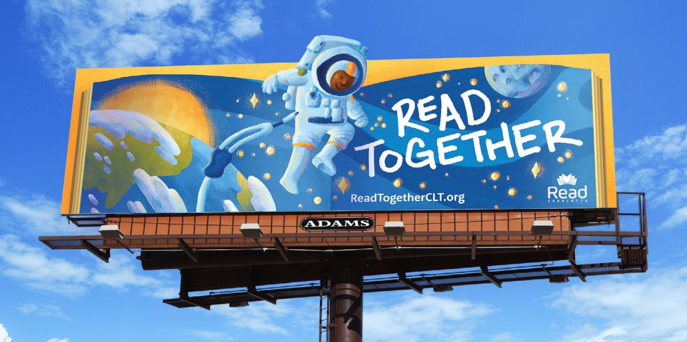 An illustrated billboard that says Read Together. It features an astronaut extending out of a book, with the Earth, Sun, and Moon behind them. ReadTogetherCLT.org