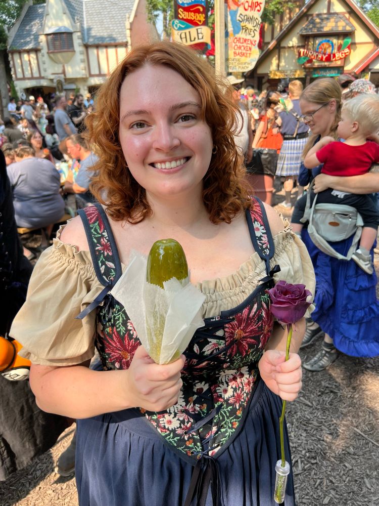 Izzy holds a purple rose in one hand and a pickle on a stick in the other hand. She smiles at the camera 