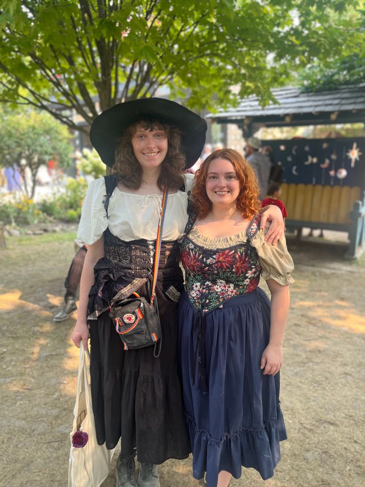 Dani and Izzy at ren faire. Dani wears a black witch hat, white top, black skirt and black corset skirt. Izzy wears a navy blue skirt, navy blue corset with flowers on it, and a light brown off the shoulder top. 