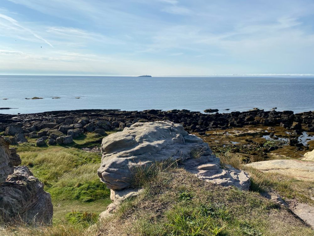 Isle of May in the Firth.of Forth. Photo taken looking out from a 
 a weathered rocky outcrop on the Fife coast.  Much of the dark rocky foreshore, visible now at low tide.  On the distant horizon about six miles out, the home of many thousands of sea bird including the amazing Puffins.  A light blue grey calm sea under a milk blue sky. The pale band on the horizon is the first signs of a  developing haar. 