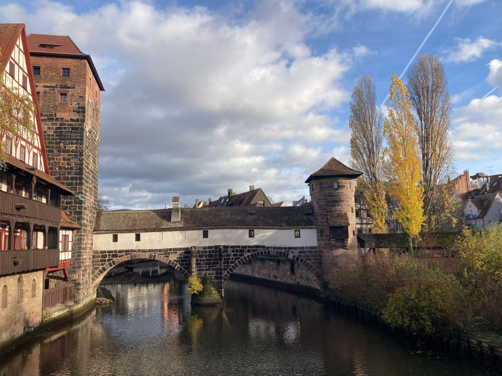 Eine Brücke mit lang gezogenem Wohnhaus dadrauf, rechts davon ein  Turm, darunter der Fluss Pegnitz. Links vorne ein Fachwerkhaus. 