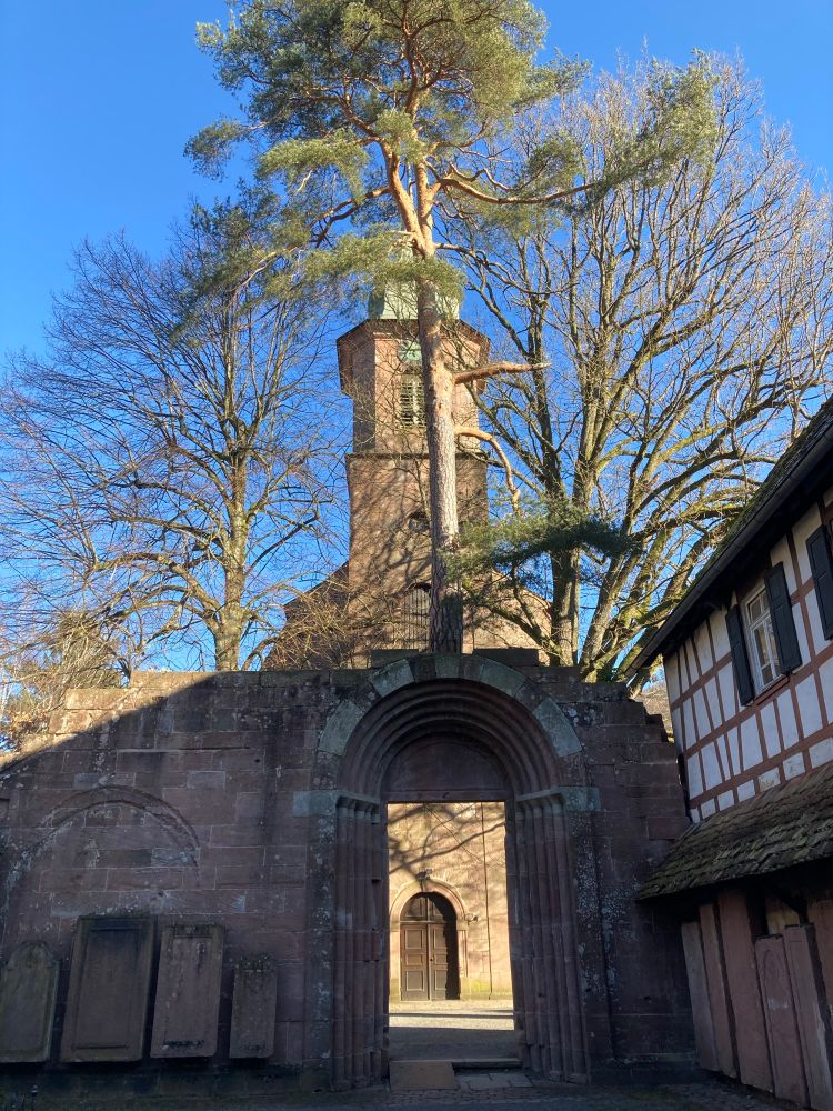 Rechts Teil eines Fachwerkhauses, daneben eine Mauer mit einem Torbogen. Auf und hinter der Mauer Bäume, dahinter eine Kirche. 