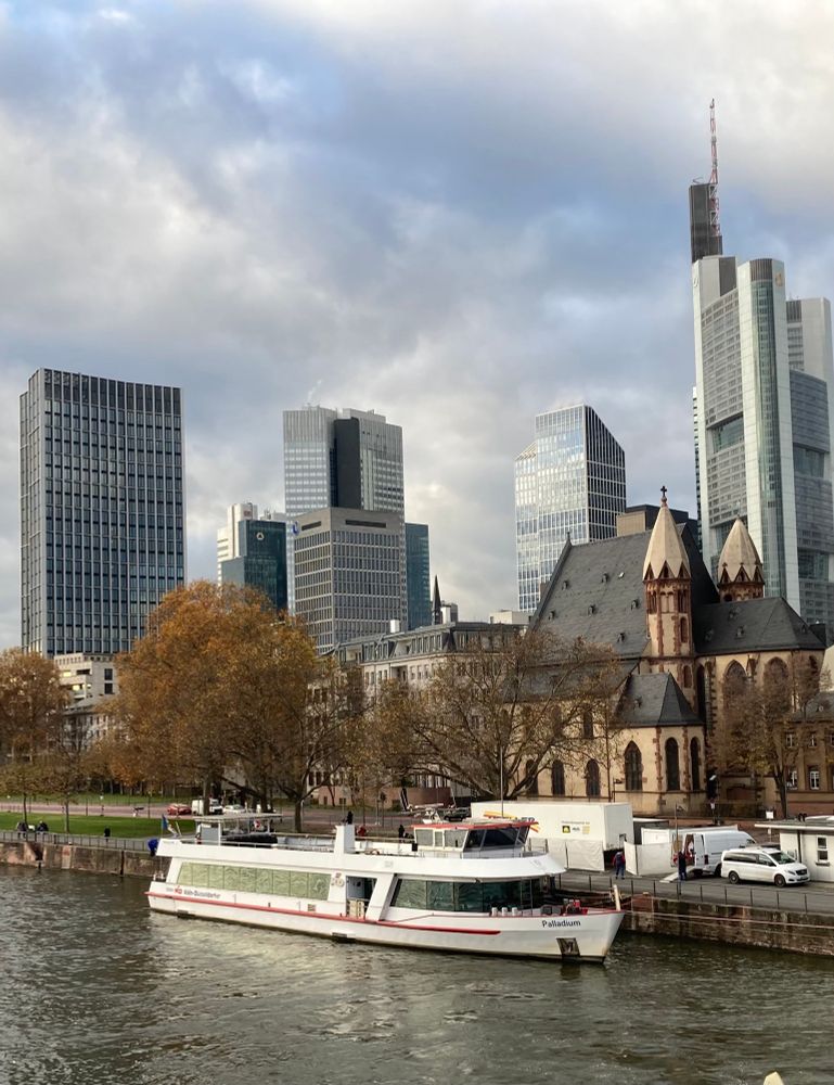 Am Mainkai in Frankfurt von der alten Brücke aus fotografiert. Man sieht den Main-Fluss, ein Schiff, Autos, die Leonhardskirche, dahinter die Hochhaus-Skyline von Frankfurt. 